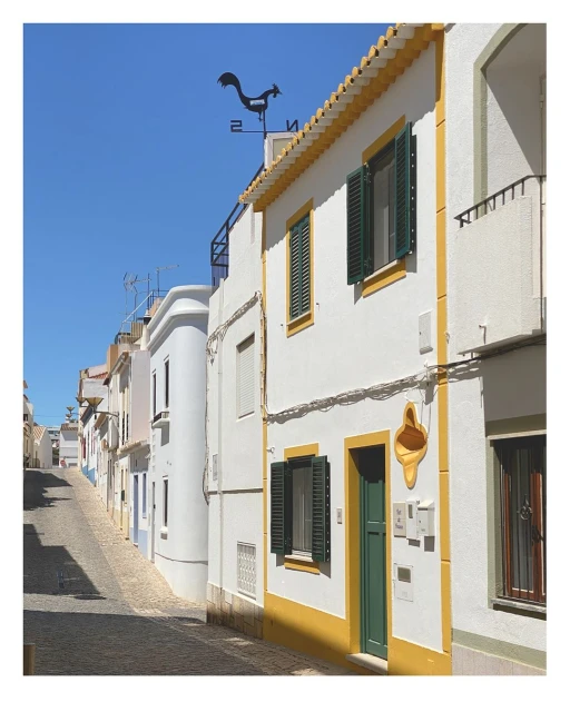 Cobblestone street lined with white buildings featuring colorful trim, shutters, and a rooftop rooster weather vane under a clear blue sky.