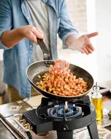 A person in a denim shirt cooks chickpeas with tomato sauce in a skillet over a portable gas burner on a tiled surface indoors.