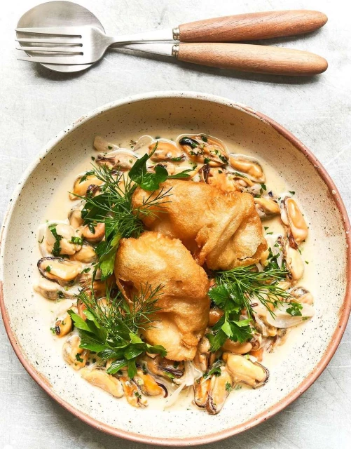 Overhead shot of a bowl of mussels and fried fish topped with dill and parsley with a serving spoon and fork on a grey surface.