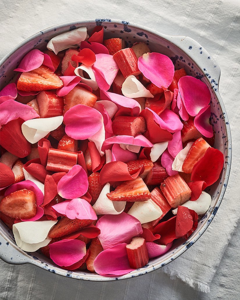 Close-up of a bowl filled with sliced strawberries and rhubarb, accented by scattered pink and white flower petals creating a colourful mix.