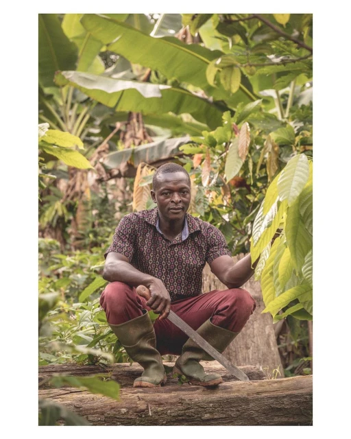 An adult African male squats on a fallen tree trunk, holding a machete, surrounded by dense green vegetation in a rural outdoor setting.