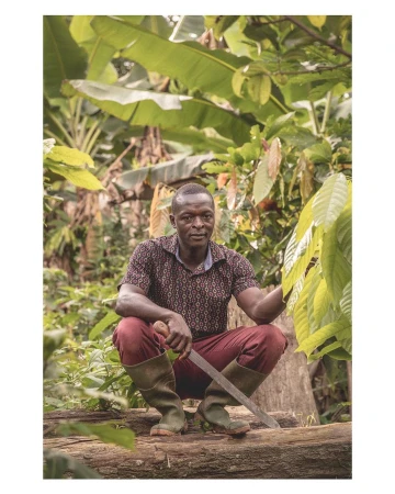 An adult African male squats on a fallen tree trunk, holding a machete, surrounded by dense green vegetation in a rural outdoor setting.
