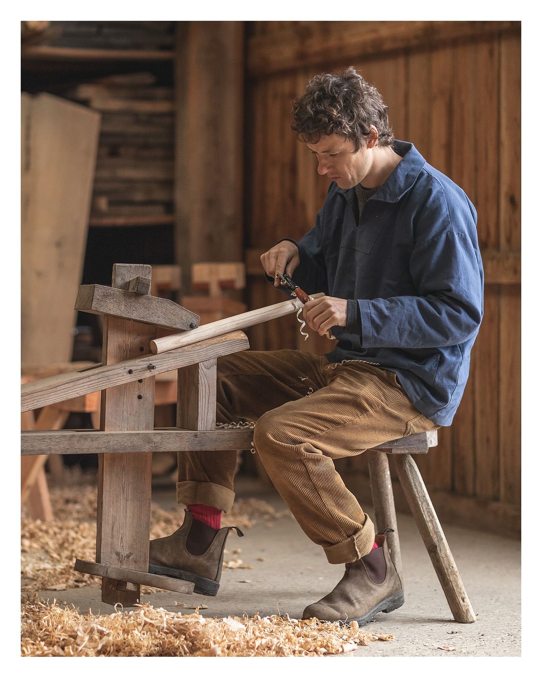 Man carves wood in a rustic workshop. Wearing a blue jacket and brown pants, he uses hand tools while seated on a wooden stool.