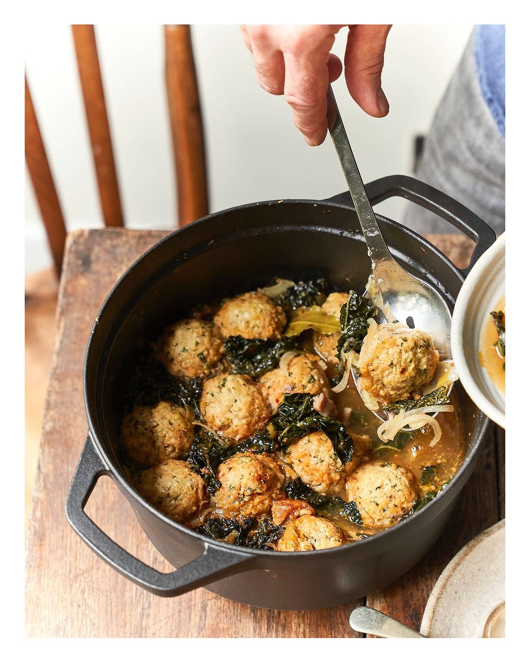 Overhead shot of a black cast-iron pot filled with meatball soup, with a hand using a slotted spoon to serve into a white bowl.