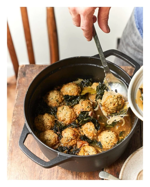 Overhead shot of a black cast-iron pot filled with meatball soup, with a hand using a slotted spoon to serve into a white bowl.