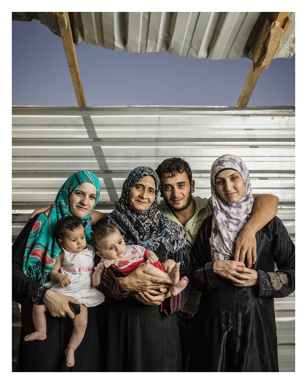 Group portrait of five Middle Eastern people with two babies in an outdoor setting with a makeshift metal wall under a blue sky.