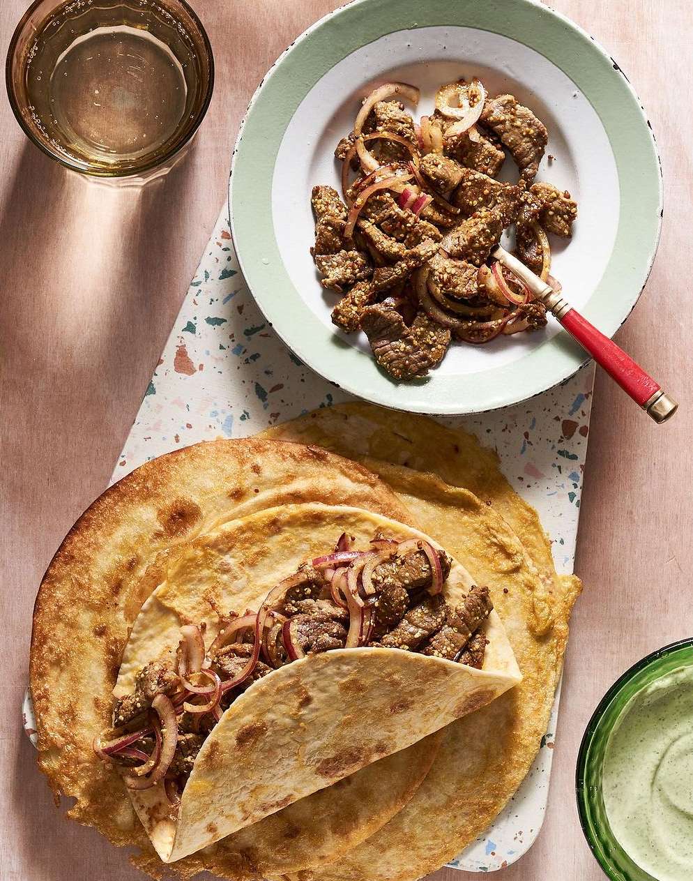 Overhead shot featuring a plate of shawarma with sliced onions, a tortilla wrap with shawarma, and a glass of clear liquid.
