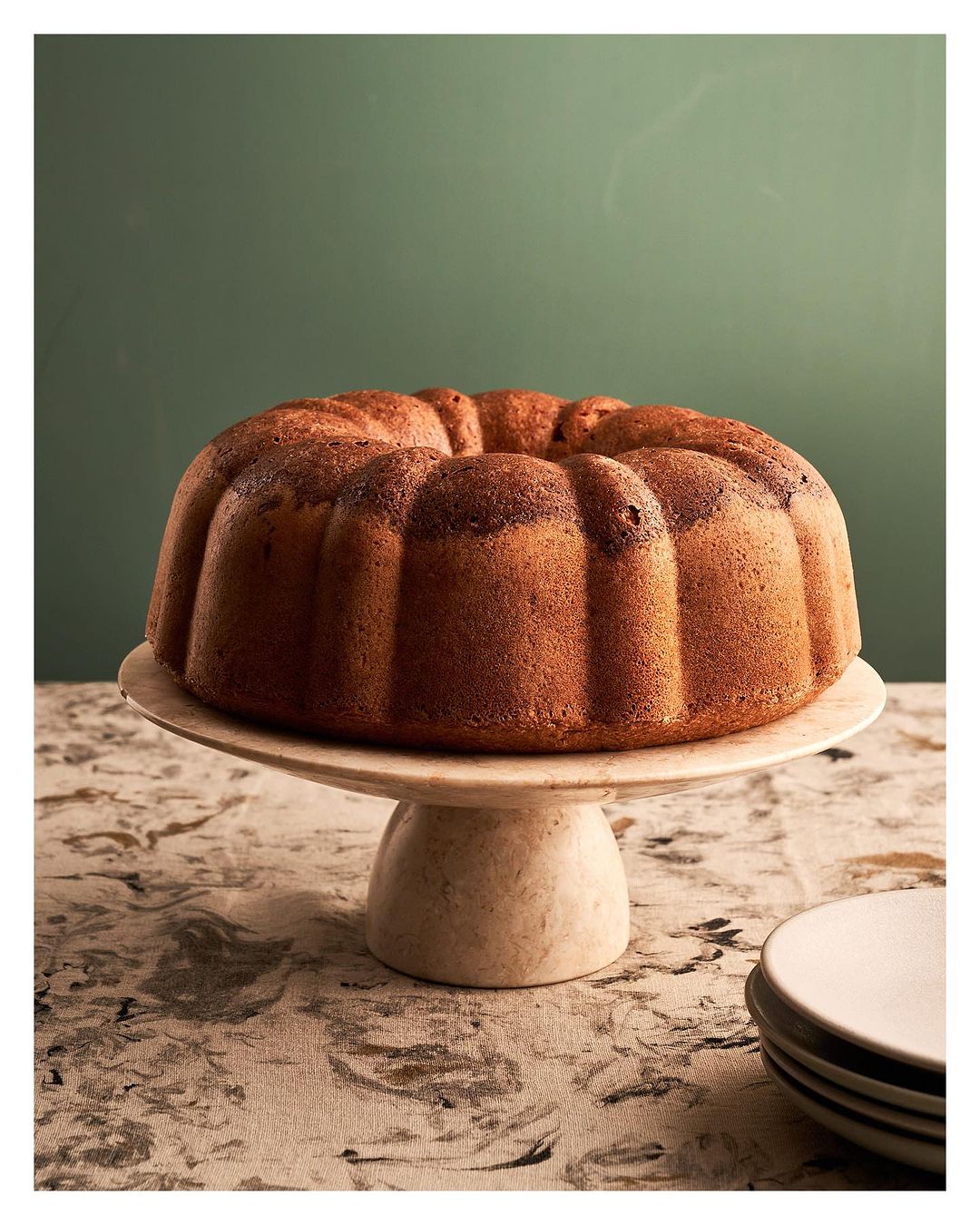 Bundt cake on a marble cake stand set on a tablecloth with a green wall in the background. Simple, elegant food photography.