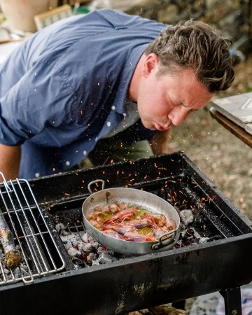 Adult male cooking seafood on a charcoal grill, blowing air into the grill; cooking outdoors with stainless steel pan with prawns.