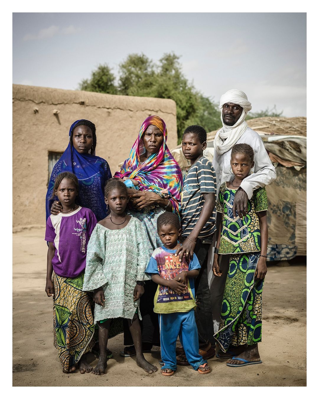 Group portrait of seven people, including adult women, men, and children, in front of a mud structure outdoors during the daytime.