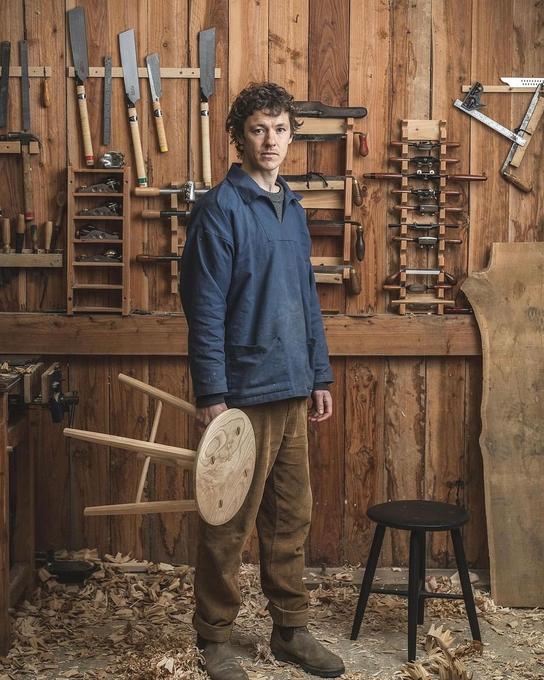 Man in workshop holding a stool with woodworking tools and wood details in the background, indoor setting and natural light.