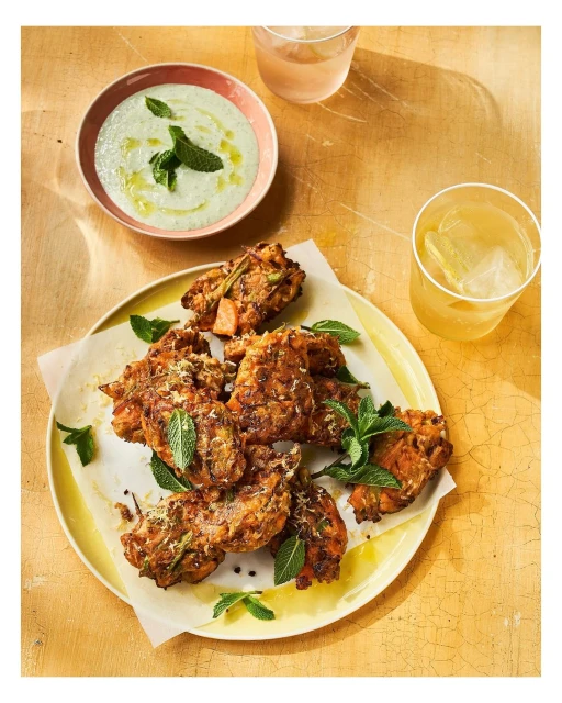 Overhead shot of a yellow plate holding crispy fried cakes garnished with fresh mint, alongside a bowl of creamy dip and drinks.