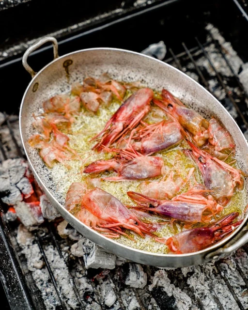Overhead shot of raw shrimp cooking in a skillet with olive oil over a charcoal grill, showcasing the vibrant colors and textures.
