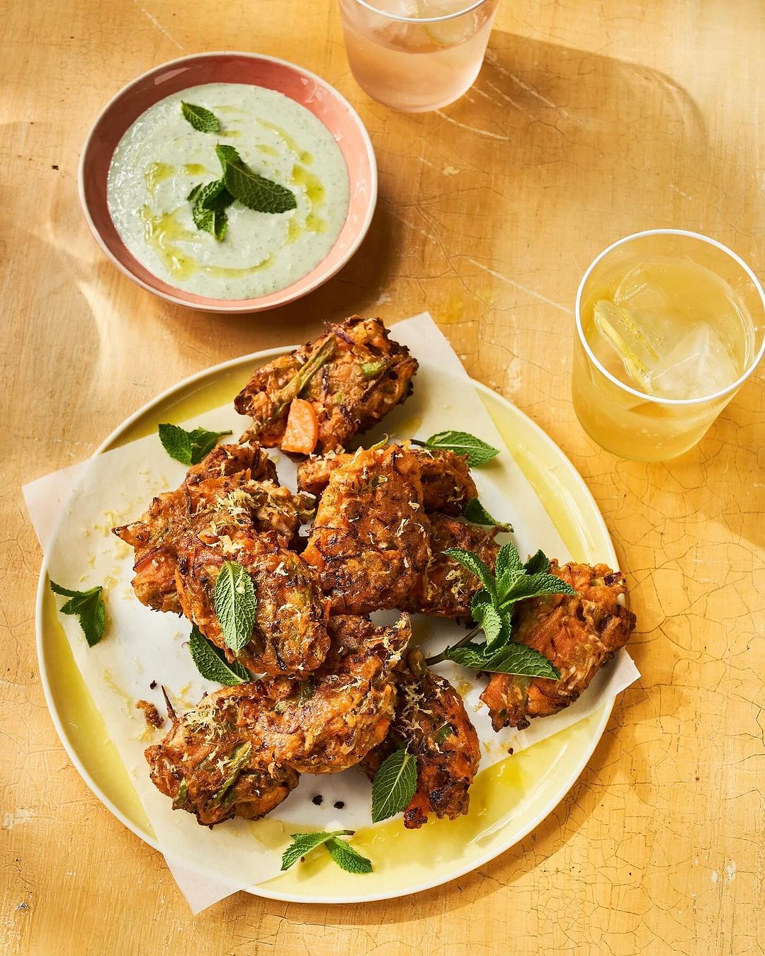 Overhead shot of fried food garnished with mint, served with yogurt sauce and drinks, presented on a yellow surface.