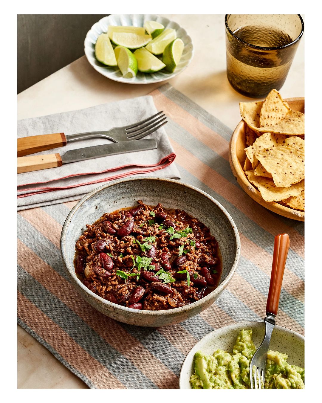 Overhead shot of chili in a stoneware bowl with lime wedges, tortilla chips, and avocado, on a striped tablecloth. Food photography.