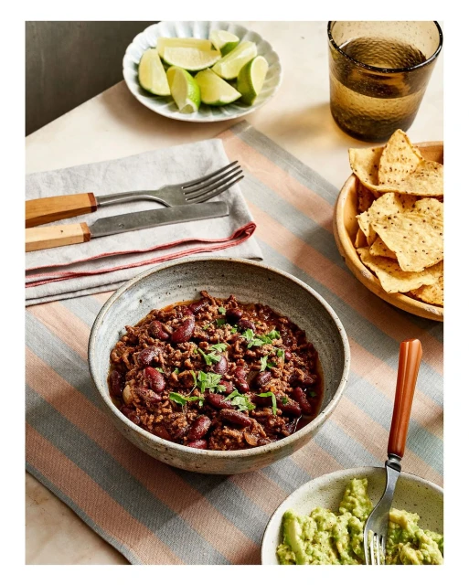 Overhead shot of chili in a stoneware bowl with lime wedges, tortilla chips, and avocado, on a striped tablecloth. Food photography.