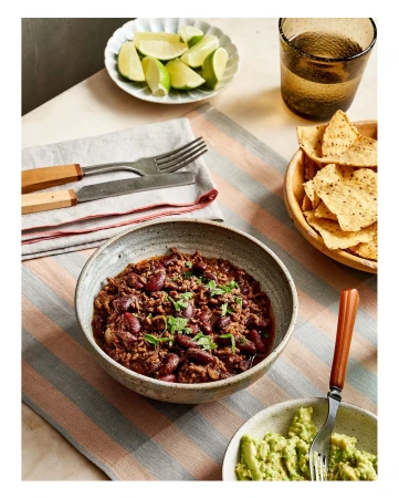 Overhead shot of chili in a stoneware bowl with lime wedges, tortilla chips, and avocado, on a striped tablecloth. Food photography.