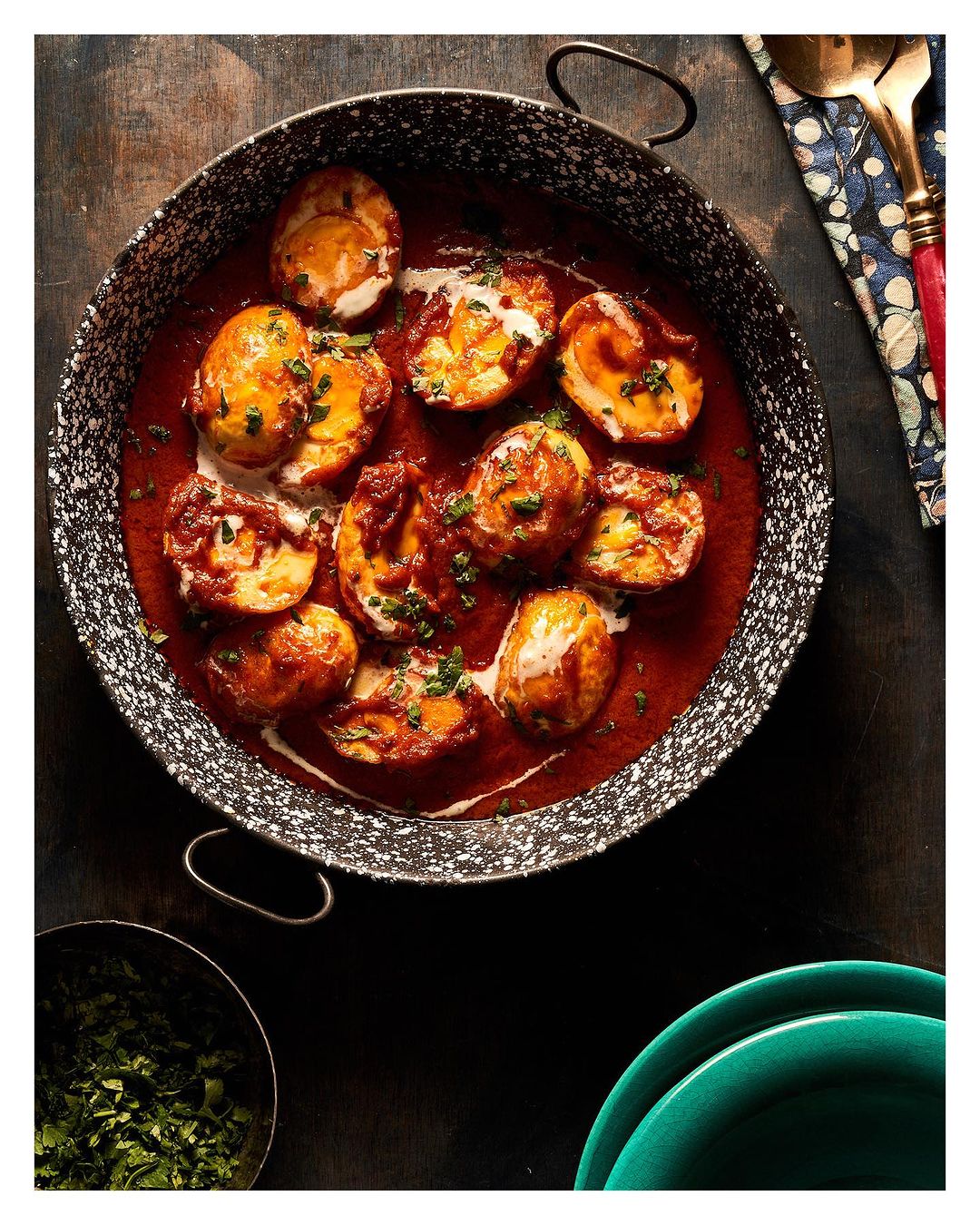 Overhead shot of an ornate pan filled with tomato-based egg curry, garnished with fresh herbs, alongside utensils and plates.
