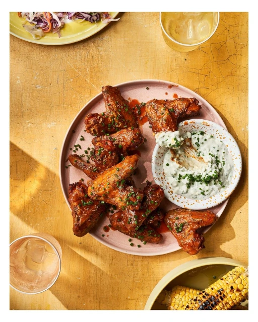 Overhead shot of glazed chicken wings with dipping sauce, cole slaw, corn on the cob, and drinks on a textured yellow surface.
