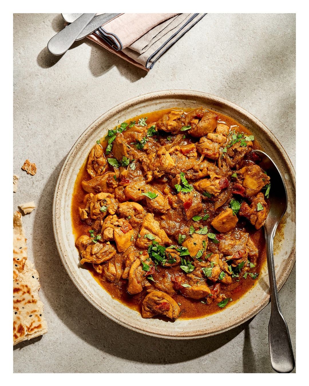 Overhead view of a plate of chicken curry with herbs, with a side of flatbread and silverware on a neutral-toned surface.