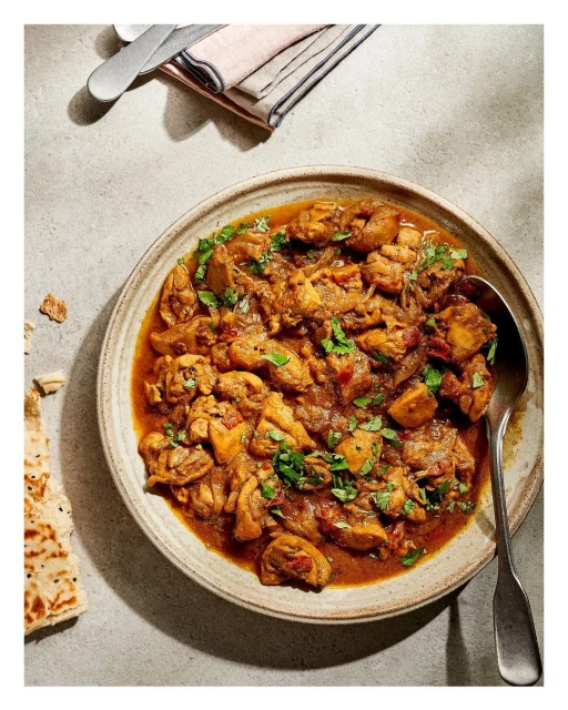 Overhead view of a plate of chicken curry with herbs, with a side of flatbread and silverware on a neutral-toned surface.