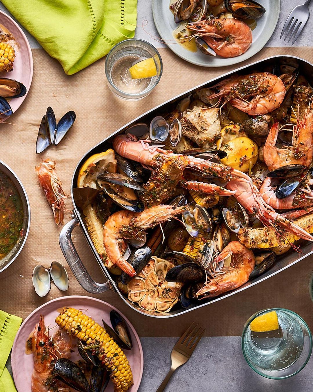 Overhead shot of a seafood boil with shrimp, mussels, corn, and clams, served in a metal tray and on plates, with drinks and napkins.