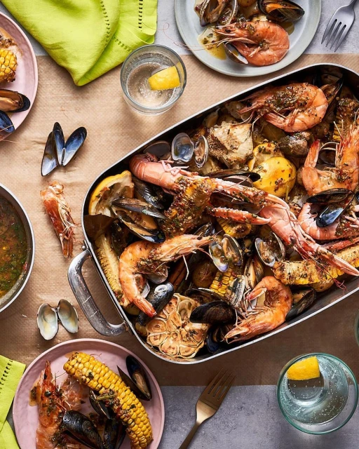 Overhead shot of a seafood boil with shrimp, mussels, corn, and clams, served in a metal tray and on plates, with drinks and napkins.