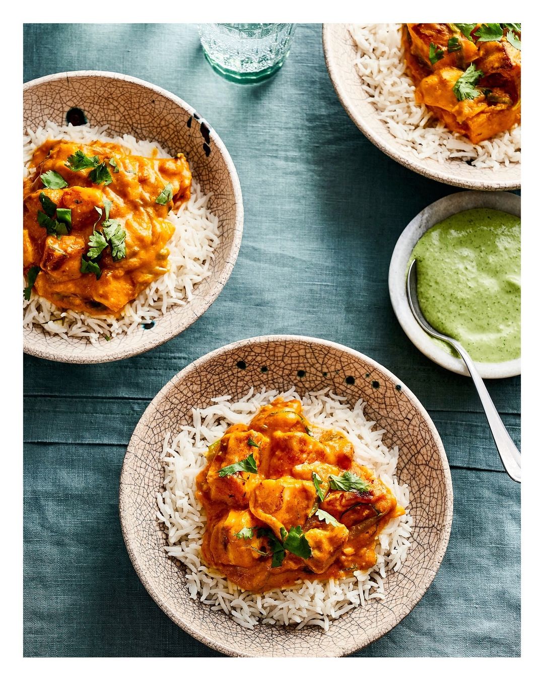 Overhead shot of three bowls with rice and sauced chicken, and a side of green sauce on a dark teal wooden table.