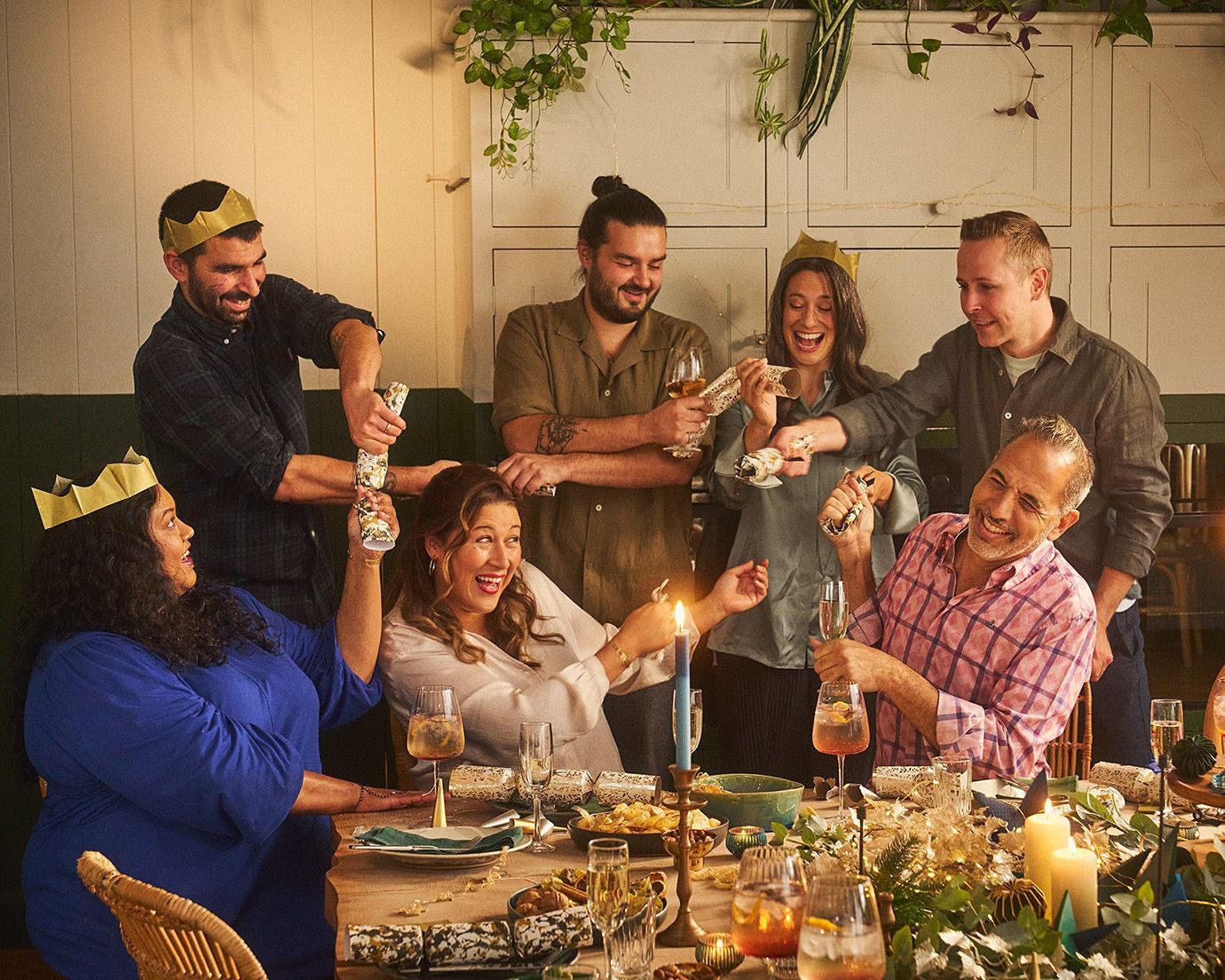 A group of adults at a dining table, pulling crackers, with the table set for a celebratory meal, and a light, warm, festive ambience.