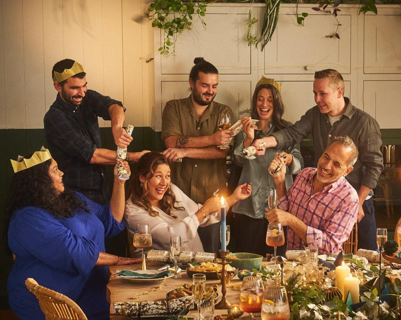A group of adults at a dining table, pulling crackers, with the table set for a celebratory meal, and a light, warm, festive ambience.
