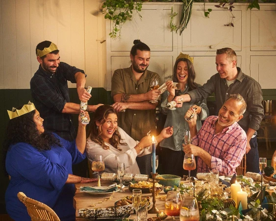 A group of adults at a dining table, pulling crackers, with the table set for a celebratory meal, and a light, warm, festive ambience.