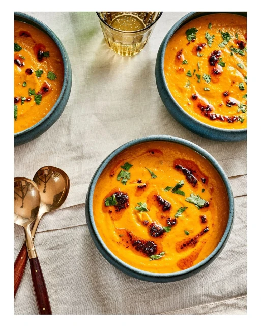 Three bowls of orange soup, garnished with herbs and chili oil, sit on a white linen tablecloth with spoons and a glass of water.