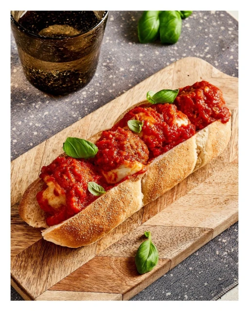 Italian meatball sub sandwich on wooden serving board with basil garnish and dark-tinted glass of beverage on grey patterned surface.