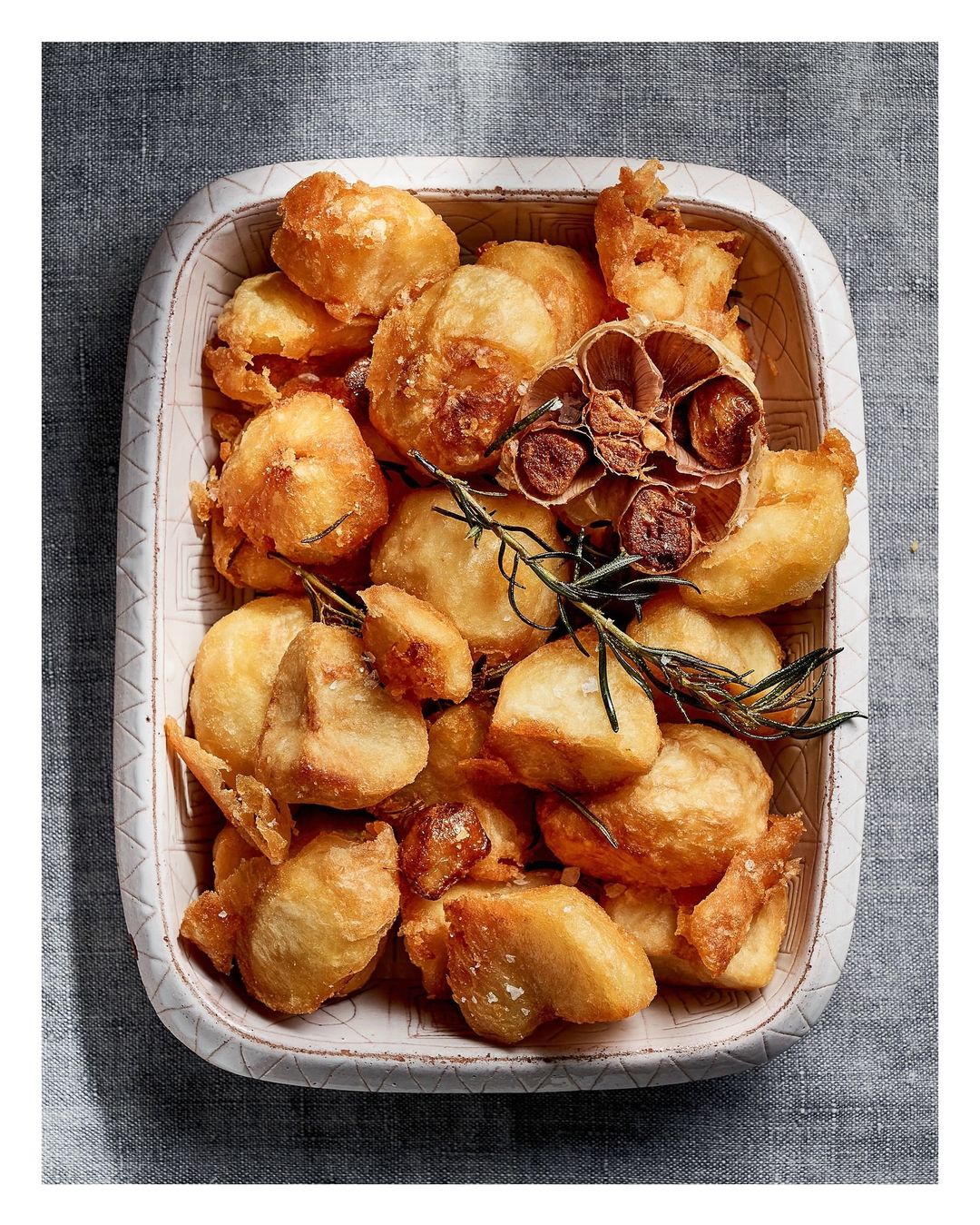Overhead shot of roasted potatoes and a bulb of garlic with rosemary sprigs in a decorative ceramic square dish on a gray linen tablecloth.