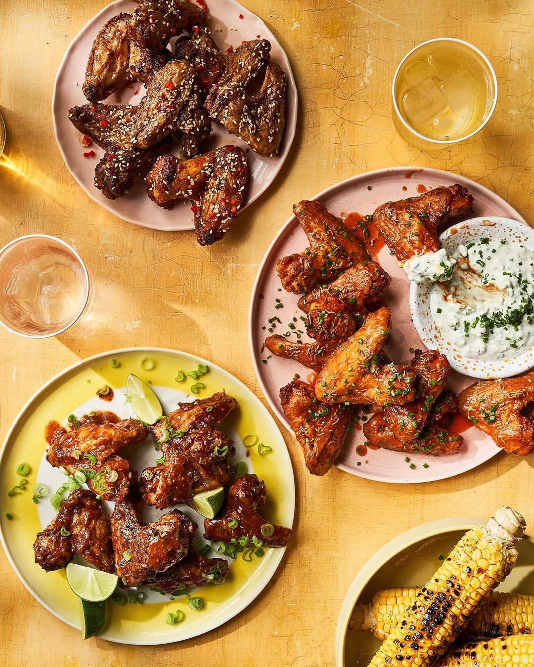 Overhead shot of three plates of chicken wings with lime wedges, garnished with green onions, served with corn and beverages.