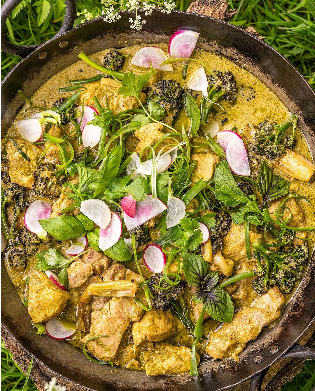 Overhead shot of a skillet filled with yellow chicken curry, garnished with broccoli florets, fresh herbs, and sliced radishes on green grass.