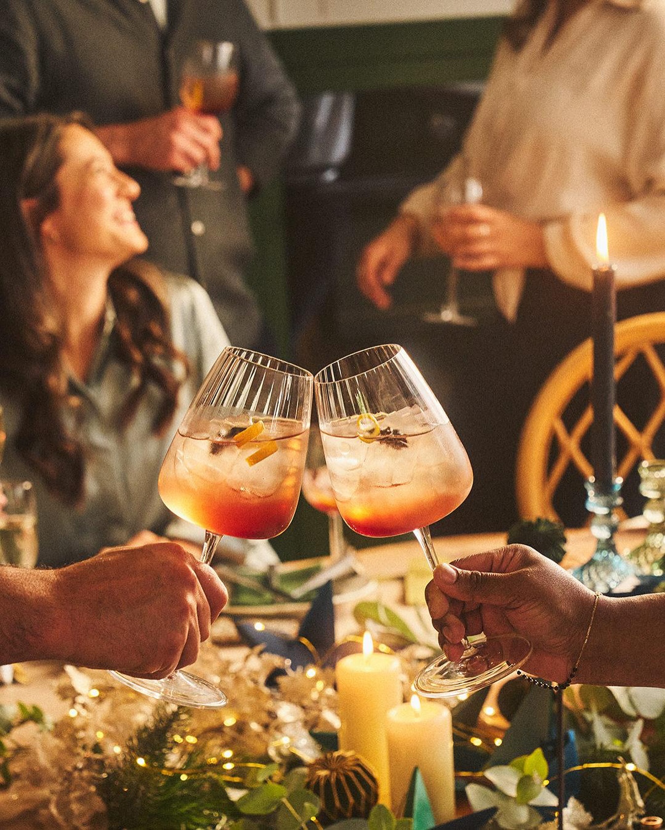 Two cocktail glasses clink together over a table decorated with garland, candles, and foliage, with several people in the background.