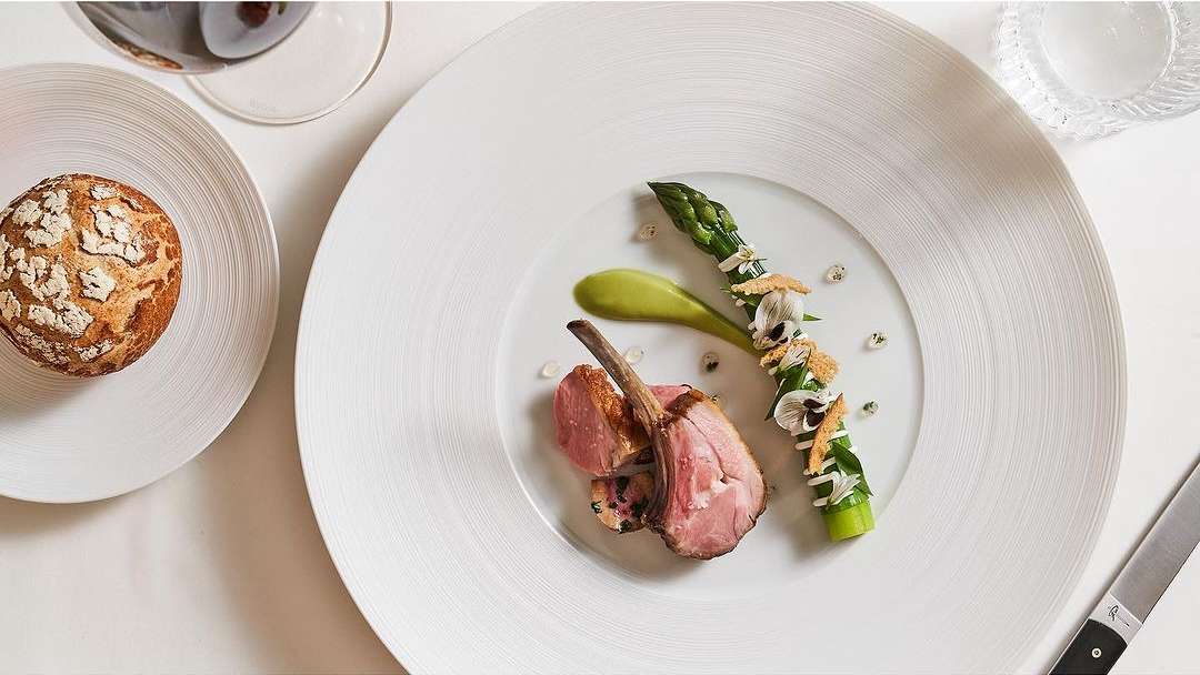 Overhead shot of a white plate with lamb chops, asparagus, green sauce, and flower garnishes, bread on side plate, and a wine glass.