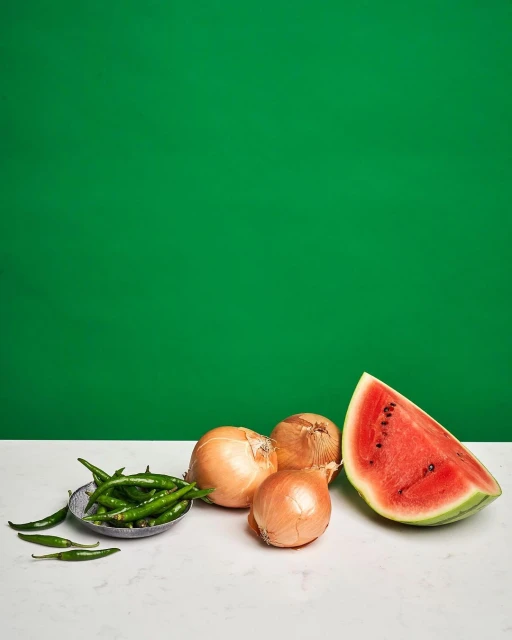 Fresh produce still life featuring a watermelon slice, onions, and green chili peppers on a marble countertop against a green backdrop.