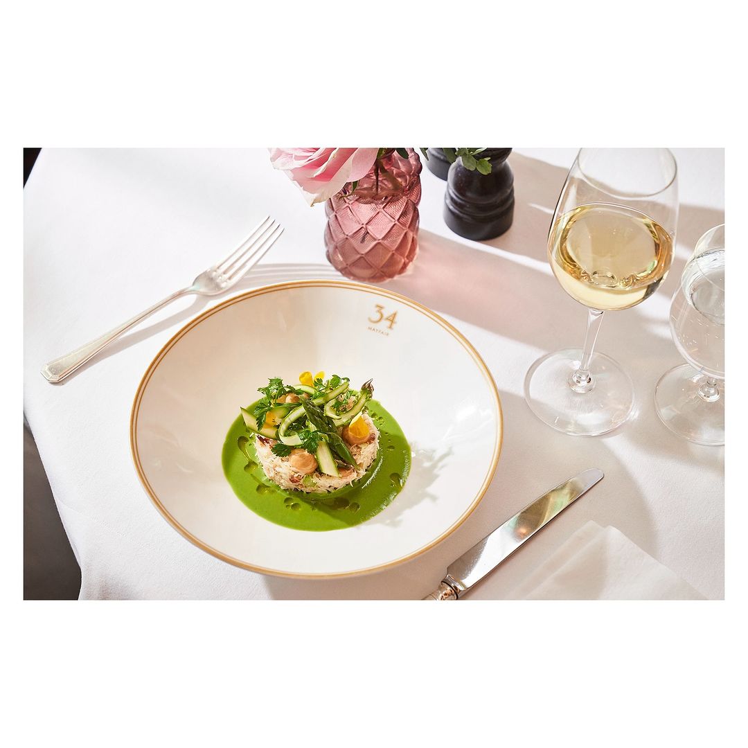 Overhead shot of a white plate with a vegetable dish, wine glass, and silverware on a white tablecloth in a restaurant setting.