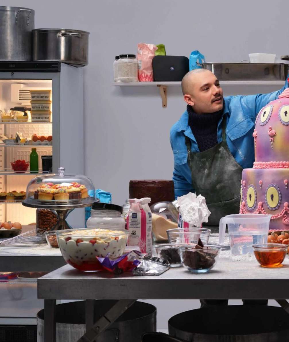 A baker decorates a whimsical cake in a commercial kitchen, surrounded by ingredients and equipment, featuring pastel colors and food industry setting.