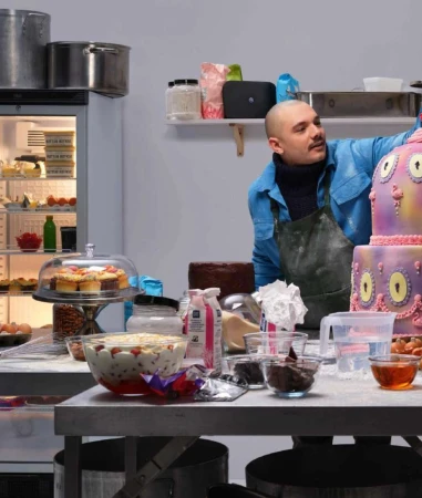 A baker decorates a whimsical cake in a commercial kitchen, surrounded by ingredients and equipment, featuring pastel colors and food industry setting.