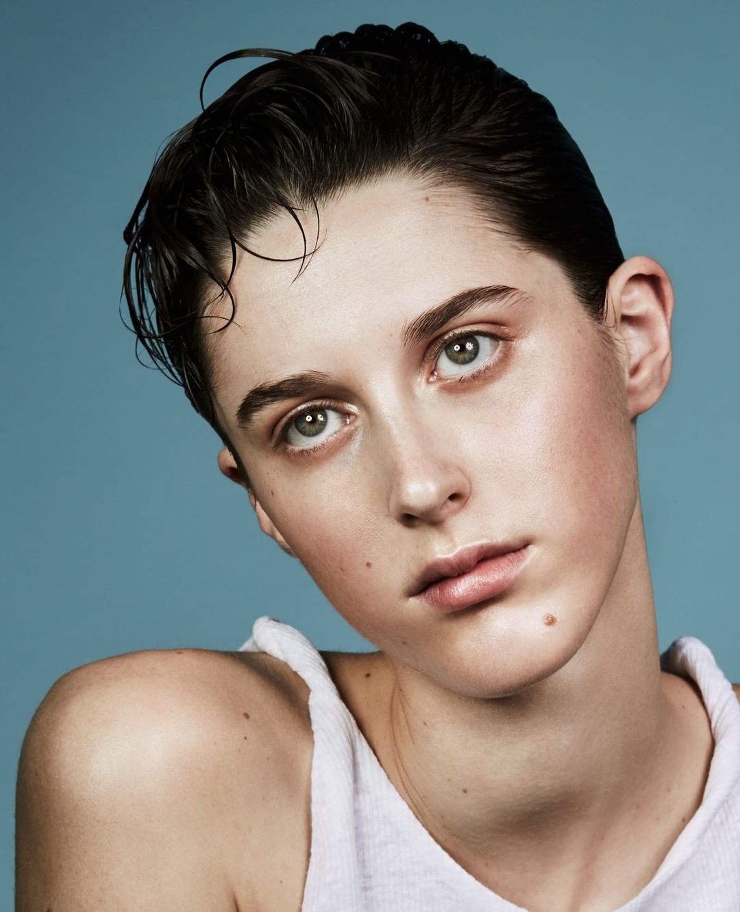 Close-up portrait of a young adult woman with wet, dark hair, light skin and a tank top against a blue background.