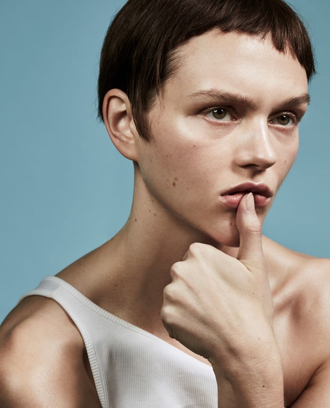 Close-up of androgynous model with finger to lips in white tank top against a light blue backdrop, showcasing a thoughtful expression.