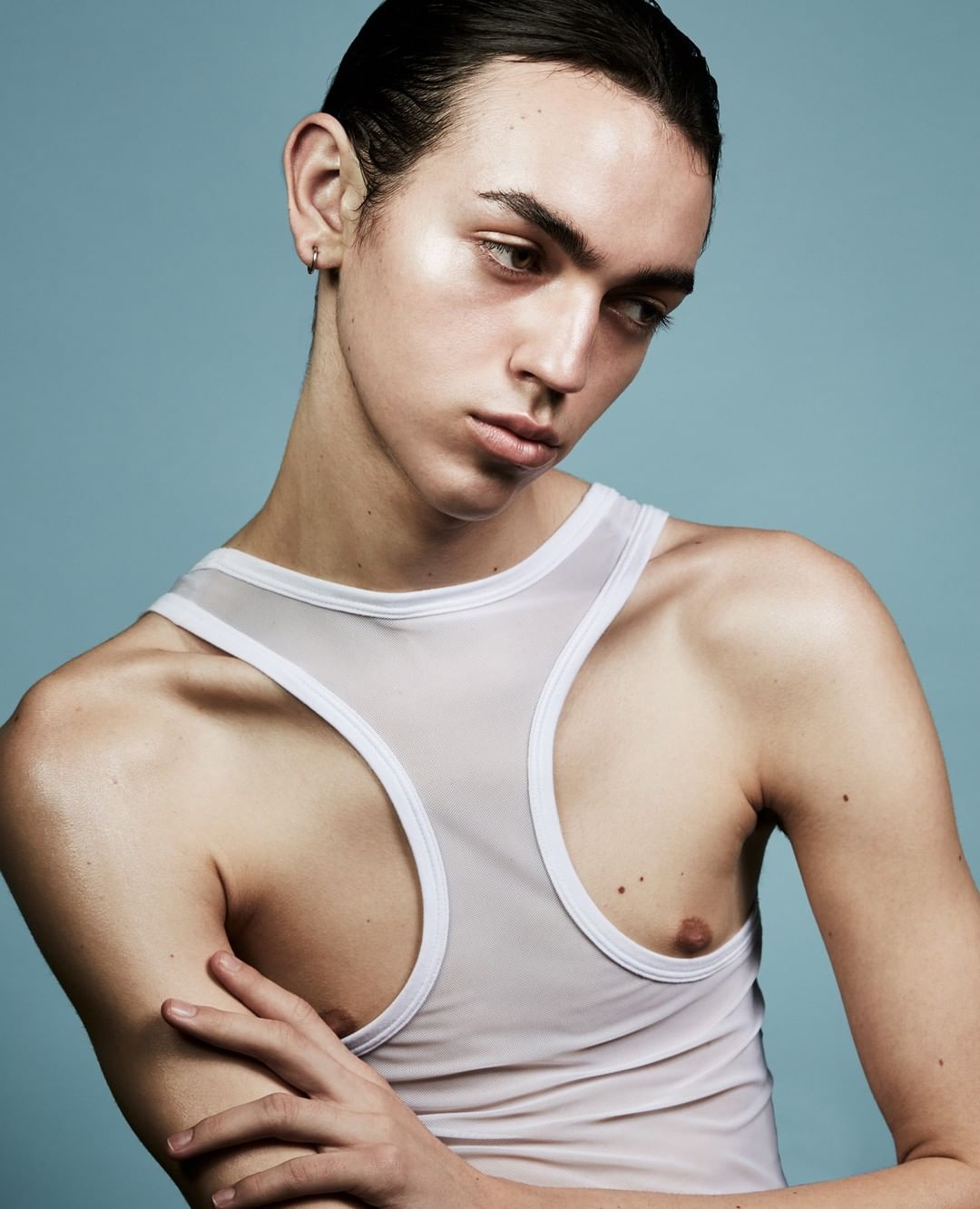 Portrait of a young adult man with slicked-back hair and a white tank top against a light blue background in a studio setting.