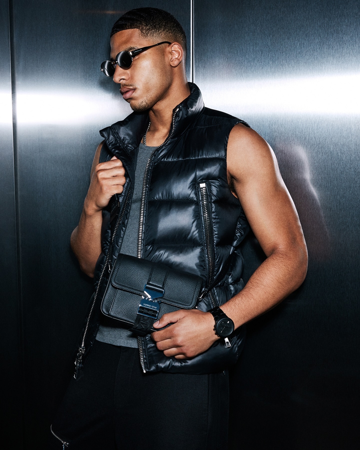 Man in puffer vest and sunglasses holds a crossbody bag in a modern setting, showcasing fashion and style.