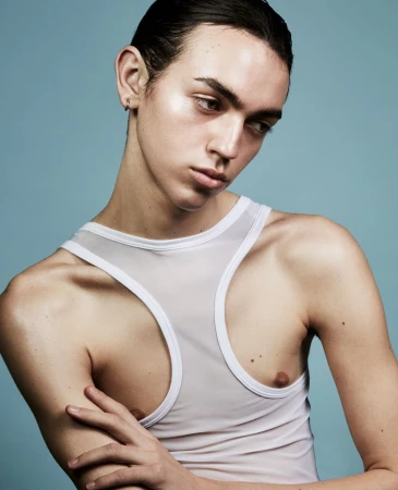 Close-up portrait of a young man with dark slicked-back hair wearing a white sheer tank top against a pale blue background.