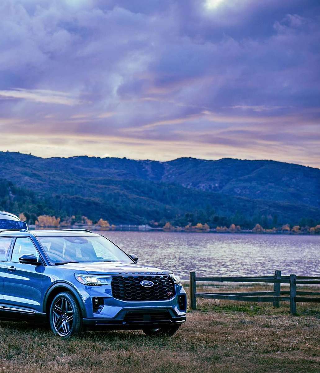 Blue Ford Explorer SUV parked on grassy field near lake and mountains under a cloudy sky at dusk, showcasing outdoor lifestyle.