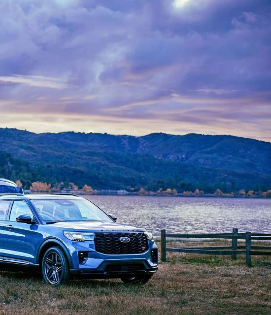 Blue Ford Explorer SUV parked on grassy field near lake and mountains under a cloudy sky at dusk, showcasing outdoor lifestyle.