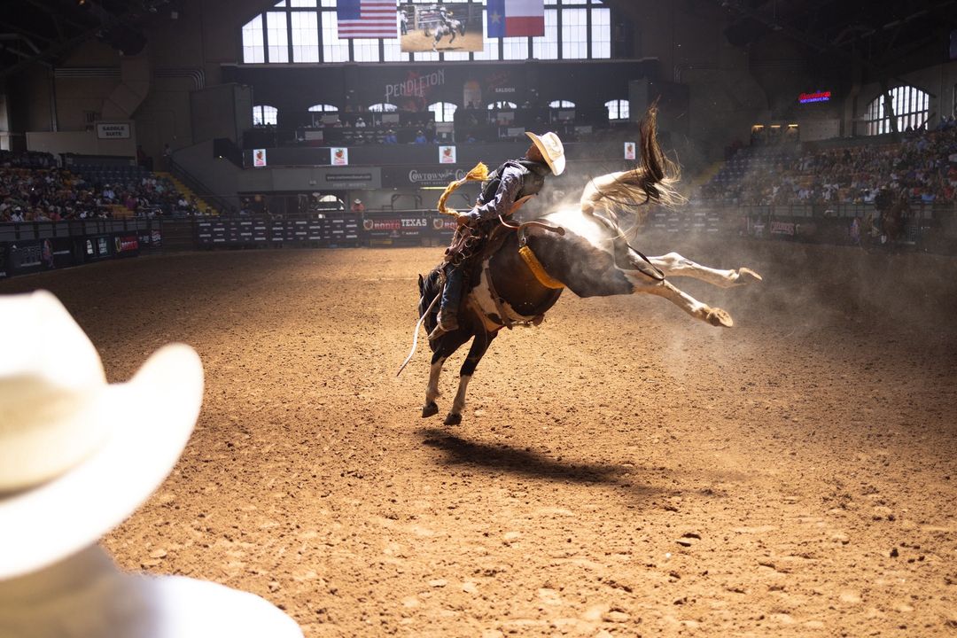 Cowboy riding a bucking bronco in a rodeo arena with a dirt ground and a crowd in the background, captured at eye-level angle.